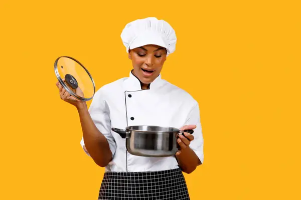 Surprised African American Chef Lady Looking Into Saucepan, Excited Black Cook Woman Holding Stewpot In Hands And Checking Meal While Cooking, Posing Isolated On Yellow Studio Background, Copy Space