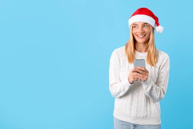 Samimi beyaz süveter giyen neşeli sarışın kadın ve Noel Baba şapkası telefonunu tutuyor. Kış yılbaşı satışlarını hevesle inceliyor, gülümsüyor, boş alana bakıyor, mavi arka plan.