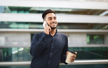 Arap girişimci cep telefonuyla iş görüşmesine daldı. Çağdaş ofis binasının yakınındaki kahve bardağının yanında duruyor. İşe verimlilik sergiliyor.