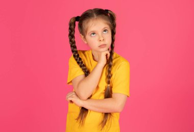 Pembe stüdyo arka planında dururken çenesine dokunan, düşüncelere dalmış, hayal kuran ya da hayal kuran, hayal kuran bir kız çocuğu, bir şeyler düşünüyor.