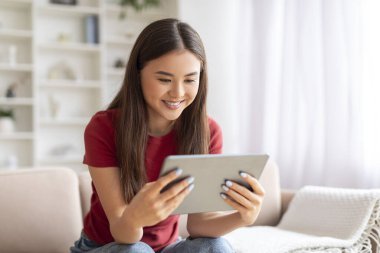 Evinde dijital tabletle rahatlayan gülümseyen Asyalı genç bir kadın. Oturma odasında dinlenirken modern aletler kullanan mutlu bir kadın. Haftasonu eğlencesinin tadını çıkarıyor.