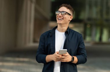 Gündelik giyinmiş, ağzı açık gözlüklü, gülümseyen, bin yıllık beyaz bir adam şehirde telefon kullanıyor, fotokopi alanına, dışarı bak. İş, iş, cihaz, serbest çalışma uygulaması, dikkat iyi haber