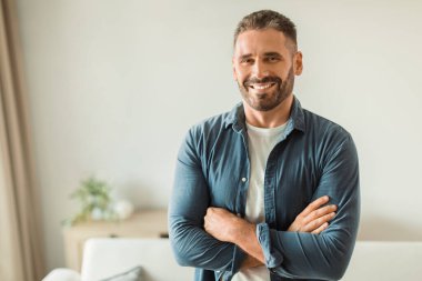 Neşeli orta yaşlı bir adam modern oturma odasının içinde ayakta duruyor. İç mekanda, kollarını kendine güvenle kavuştururken gülümsüyor, gündelik kıyafetlerle poz veriyor ve olumlu duygularını ifade ediyor.