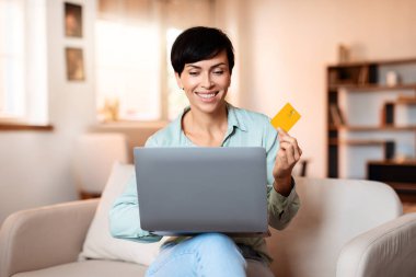 Orta yaşlı mutlu bir kadın dizüstü bilgisayardaki mağazaları karıştırıyor, kredi kartıyla ödeme yapıyor, oturma odasındaki kanepede oturuyor. İnternet alışverişi dijital kolaylıkları