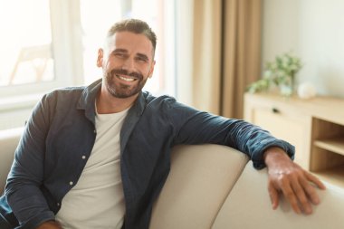 Evdeki rahat koltukta oturan mutlu orta yaşlı adam, dostça gülümseyerek kameraya bakıyor, modern oturma odasındaki hoş ve hoş atmosferin tadını çıkarıyor.