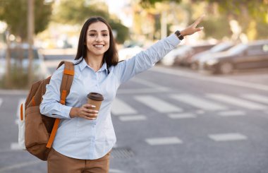 Sırt çantasıyla gülümseyen genç Avrupalı bir kadın, bir fincan kahve servisi, açık havada el işaretli bir taksi durağı. Bayan seyahatin, turistin, reklamın ve teklifin tadını çıkar.