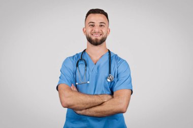 Mavi önlüklü ve steteskoplu Avrupalı erkek doktor, kollarını kavuşturmuş, tıbbi yardım ve bakım sağlamaya hazır, profesyonellik ve uzmanlık gösteriyor.