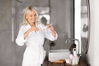 Güzel son sınıf öğrencisi dişlerini diş fırçasıyla fırçalıyor, mutlu olgun bir kadın aynanın yanında duruyor, sabah temizliğini evde yapıyor, ağız bakımı yapıyor, uzay kopyaları yapıyor.