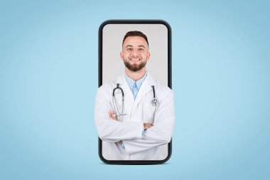 Genç gülümseyen Avrupalı erkek doktor kollarını kavuşturmuş, steteskop boynuna dolanmış, mavi arka planlı cep telefonu ekranında poz veriyor.