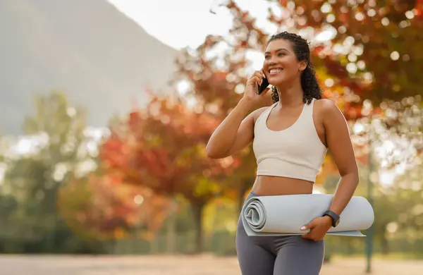 Happy sporty woman calling on cellphone standing with fitness mat, ready for workout exercise in nature, posing in park outdoors, talking by phone and looking aside. Copy space