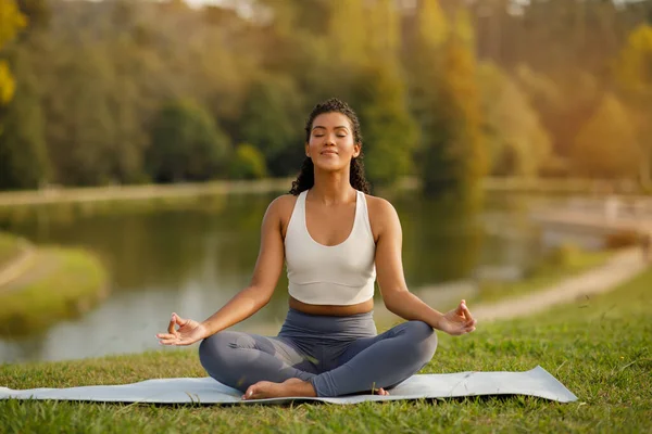 Young fitness woman in fitwear practices morning yoga sitting in lotus position, meditating on mat in nature near lake in green park outdoors. Lady enjoys calmness during workout meditation