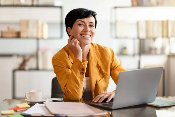 Middle aged business lady at laptop posing for portrait at workplace, smiling to camera while browsing online website, sitting at desk in modern office interior. Professionalism and technology