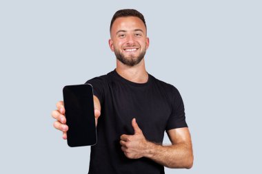 Sakallı güçlü beyaz bir adam, ekranı boş bir akıllı telefon ve gri stüdyo arka planında izole edilmiş el hareketleriyle hareket ediyor. Spor onaylandı, vücut bakımı tavsiyesi