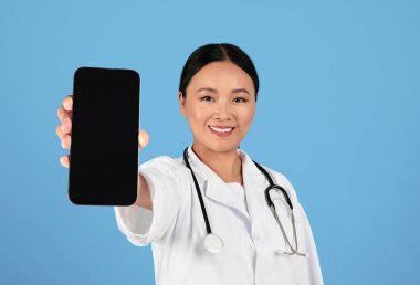 Mavi Stüdyo 'nun geçmişine poz verirken elinde siyah ekranla Büyük Boş Akıllı Telefonu olan Mutlu Asyalı Doktor Kadın Sağlık Sigortası veya Tıbbi Uygulama Reklamcısı