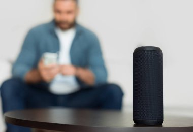 Kablosuz akıllı hoparlör masanın üzerinde dururken tanınmayan adam, müzik listesine bakıp Modern Oturma Odasında Oturup Dinleniyor. Seçici Odaklanma