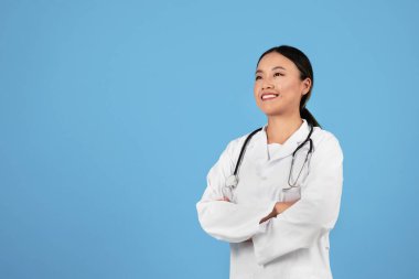 Tıbbi Hizmetler. Kendine güvenen Asyalı doktor üniformalı kadın, kolları katlanmış, mavi stüdyo arka planında, beyaz önlüklü ve steteskoplu kadın, başka tarafa bakıyor ve gülümsüyor, boşluğu kopyalıyor.