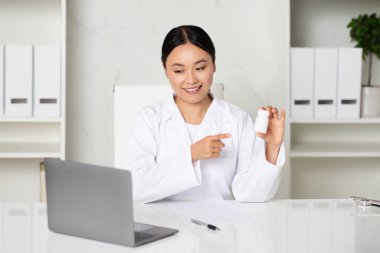 Gülümseyen Asyalı doktor kadın, kliniğin içinde dizüstü bilgisayarla video çekerken ilaç kavanozu gösteriyor. Doktor kadın hastayla telekonferans yapıyor, internetten danışmanlık yapıyor, tedavi öneriyor.