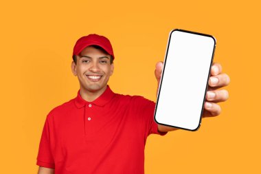 Üniformalı bir Orta Doğulu kurye, şablon için boş cep telefonu ekranı sunuyor, sarı arka plandaki stüdyoda online alım ve teslimat uygulaması öneriyor. Model