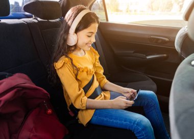 Kulaklıklı neşeli genç kız arabaların arka koltuğunda otururken akıllı telefonunun keyfini çıkarıyor. Güneş ışığı içeri giriyor, yanında kırmızı okul çantası var.