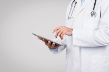 Beyaz önlüklü ve steteskoplu erkek doktor, gri arka planda sağlık hizmetlerinde teknolojik bütünleşmeyi gösteren dijital tablet, navigasyon veya girdi bilgileriyle ilgilenmektedir.