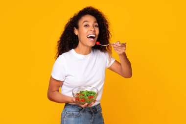 Genç siyah kadın yeşil salatanın tadını çıkarıyor, sarı stüdyo arka planında gülümsüyor. Sağlıklı yaşam tarzını ve beslenme tarzını benimseyen, sağlıklı beslenme düzenini zayıflatan bir kadın.