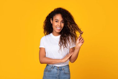 Kıvırcık saçlarına dokunan, sarı stüdyo arka planında kameraya güvenle gülümseyen genç Afrikalı Amerikalı kadın. Doğal güzelliği ve saç bakımını kucaklamak