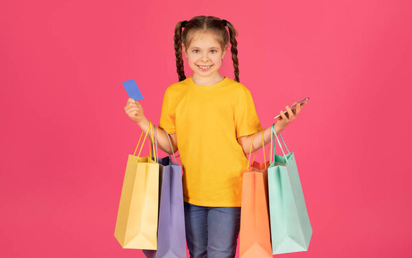 Great App. Счастливая маленькая девочка, держа в руках сумки с покупками и кредитные карточки, милая дочка, получающая удовольствие от совершения онлайн-покупок в мобильном приложении, стоящая на розовом фоне студии