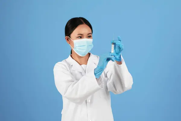 Vaccination Concept. Asian doctor woman in protective medical mask looking at syringe in her hand, physician lady wearing uniform standing isolated over blue background, ready to make injection