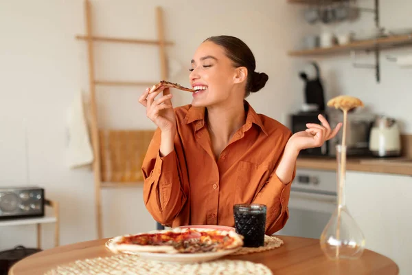 Cheerful young woman in orange blouse enjoys bite from pizza slice, seated in modern kitchen with ambient lighting and minimalist decor, free space