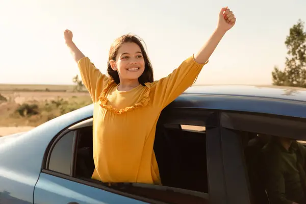 Happy girl joyfully raises her arms out of blue car, feeling the freedom of the open road on sunlit day, with vast landscape in the background