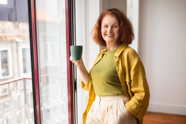 Happy Senior Woman Drinking Tea While Standing Near Window At Home, Smiling Beautiful Elderly Woman With Cup Of Hot Drink Relaxing Indoors, Enjoying Domestic Comfort And Retirement Leisure