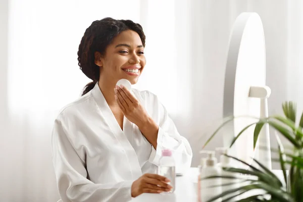Skin Cleansing. Beautiful Black Woman Using Micellar Water And Cotton Pad At Home, Attractive African American Lady Sitting At Dressing Table, Making Daily Skincare Routine, Copy Space