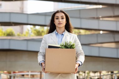 Ciddi, genç, beyaz kadın yöneticinin elinde ofis malzemeleriyle dolu karton kutu, yeşil saksı bitkisi, şehir ofisinde, açık havada. İşle ilgili sorunlar, işten çıkarma, istifa