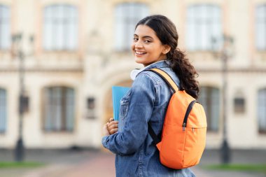 Akademik kitapları ve çantasıyla, eğitim kurumunun önünde poz veren ışıl ışıl genç bir Hintli akademisyen lense sıcak bir gülümseme sunuyor. Başlık için alan, eğitimin vurgulanması.