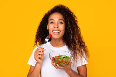 Afrikalı Amerikalı bir kadın, stüdyoda sarı arka planda kameraya gülümseyen sebze salatasının tadını çıkarıyor. Sağlıklı yemek ve çatalla poz veren kadın portresi. Dengeli yemek seçmenin güzelliği