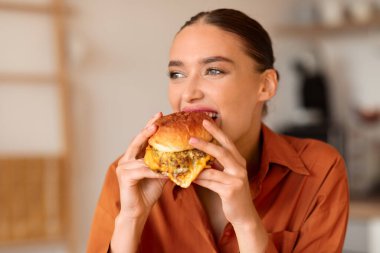 Neşeli kadın yeni yapılmış hamburgerin tadını çıkarıyor, arka planda ince detaylarla ev mutfağının arka planına kurulmuş.