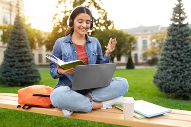 Neşeli genç Hintli bir kız öğrenci, ıssız bir üniversite kampüsünde, bilgisayarını ve kulaklıklarını kullanarak, muhtemelen online bir derse ya da seminere katılıyor. E-eğitim