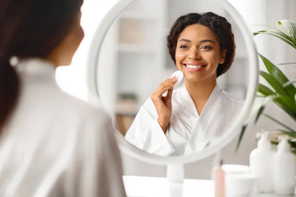 Beauty Routine. Smiling Black Woman Using Cotton Pad For Cleansing Face At Home, Happy African American Lady Looking In Mirror, Enjoying Domestic Skincare Treatments, Selective Focus On Reflection
