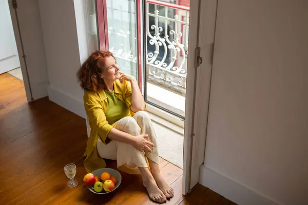 自宅や居心地の良いホテルの部屋の近くの床に座っている成熟した赤毛の女性 グラスワインと果物のプレートでリラックスして笑顔のシニア女性 退職時に彼女の時間を楽しんで スペースをコピー — ストック写真