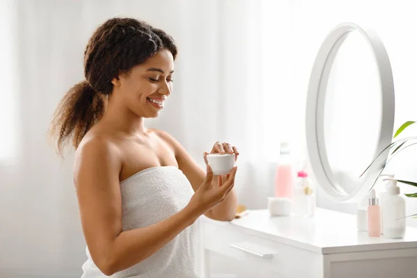 Cosmetics Concept. Beautiful African American Female Using Moisturising Cream While Sitting Near Mirror At Home, Happy Black Woman Opening Jar With Nourishing Lotion, Making Beauty Skincare Routine