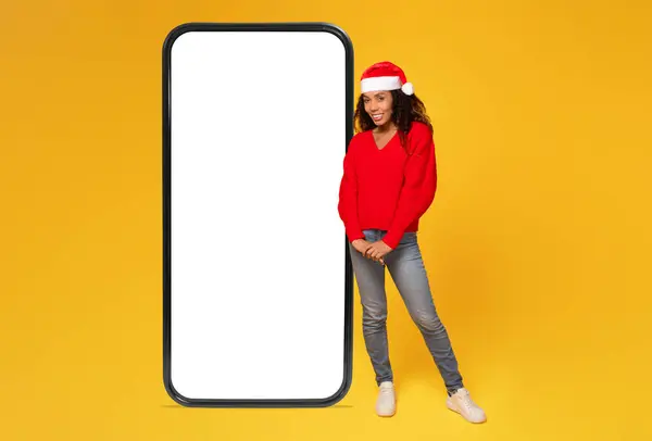 Happy black lady in Santa hat stands by giant phone with white blank display, yellow background, ideal for Christmas promos, full length, free space