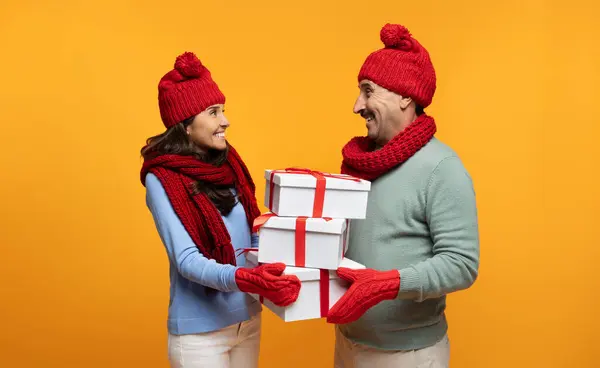 Happy senior european wife in hat give many boxes gifts to husband at winter, isolated on orange studio background. Relationships, present for New Year, Christmas celebration