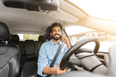 Mutlu, sakallı, genç Hintli adam araba sürerken telefonla konuşuyor, güneş parlıyor. Neşeli doğulu adam içeride oturuyor, telefonla konuşuyor.