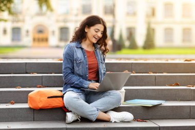 Gülümseyen Hintli kız öğrenci merdivenlerde oturuyor dizüstü bilgisayarla çalışıyor açık havada sınavlara hazırlanıyor, üniversite kampüsünde dinleniyor. Teknoloji, eğitim ve uzaktan çalışma kavramı, kopyalama alanı