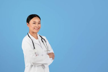 Sağlık Hizmetleri Profesyoneli. Beyaz laboratuar önlüklü, kolları çapraz duran, gülümseyen, mavi stüdyo arka planına karşı duran, steteskoplu, neşeli Asyalı kadın doktor.