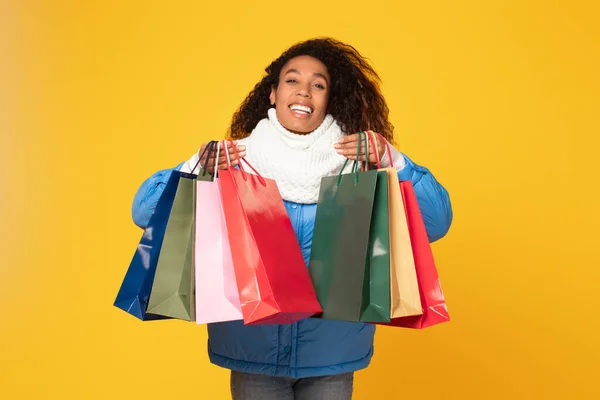 Excited black woman in winter attire is gleefully presenting an array of vibrant shopping bags, illuminating the warm, yellow background with a sense of retail triumph