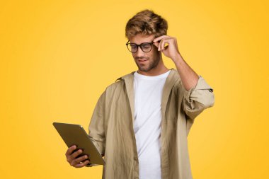 Tarz sahibi Avrupalı adam sarı stüdyo arka planında duruyor, dijital tableti tutuyor ve gözlüklerine nazikçe dokunuyor, uygulama veya sörf sitesi kullanıyor.