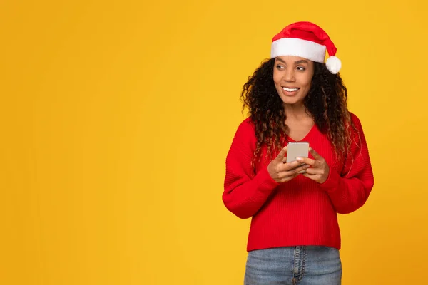 Happy young black lady in Santa hat holding cellphone and looking aside at free space against yellow background, embodying Christmas offers