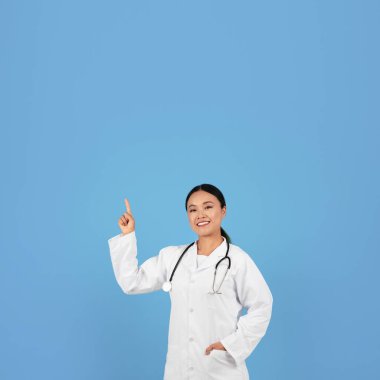 Gülümseyen Asyalı Kadın Doktor Mavi Stüdyo Arkaplanı 'nın üzerinde dururken Başının üzerindeki Kopya Uzayı' nı işaret eden, Doktor Üniformalı Kadın Tıbbi Reklam İçin Boş Yer Gösteriyor