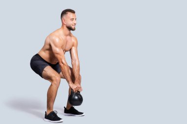 Mutlu terli, güçlü, kaslı, sakallı, çıplak gövdeli beyaz adam gri stüdyo arka planında izole edilmiş kettlebell ile egzersiz yapıyor. Vücut bakımı, güç egzersizi, spor